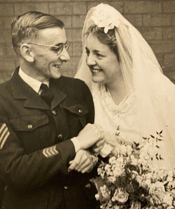 Sandy Hinds in military uniform and Coral Osbourne in a wedding dress hold hands and smile at one another.