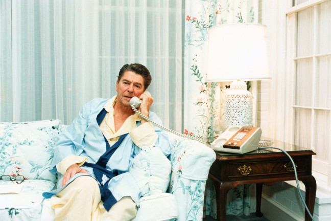 Ronald Reagan sitting on a coach in pyjamas and a robe, speaking on a phone.