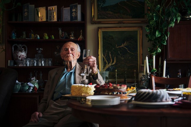 Captain Victor Lederer seated at a table with multiple cakes on it. He raises a glass in his left hand. 