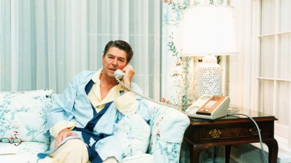 Ronald Reagan sitting on a coach in pyjamas and a robe, speaking on a phone.